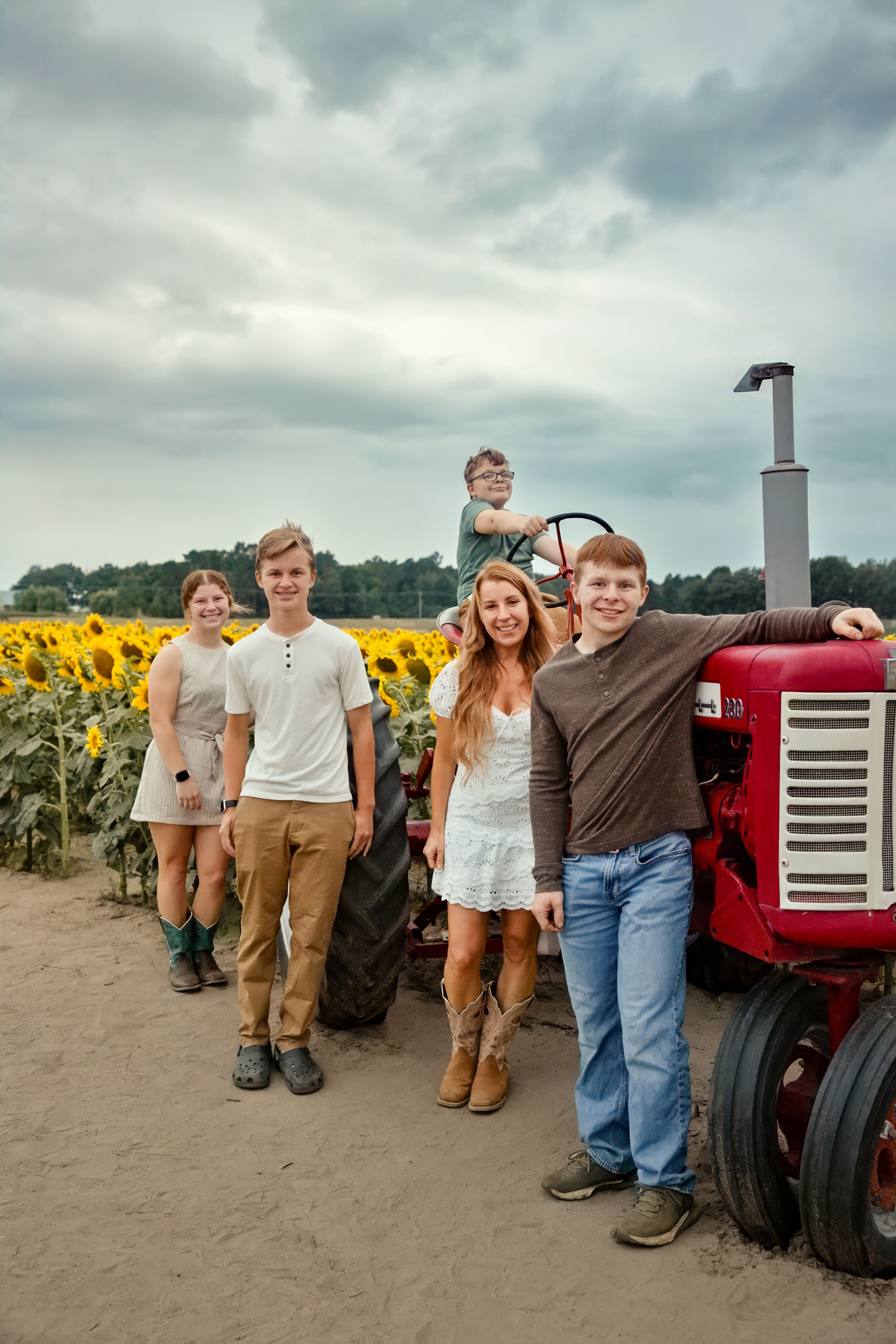 Family of five posing near a red tractor beside a sunflower field on a cloudy day.