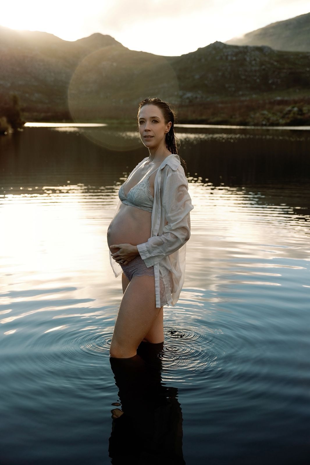 Pregnant woman standing in a calm lake at sunset, wearing a bikini and open shirt, cradling her baby bump.