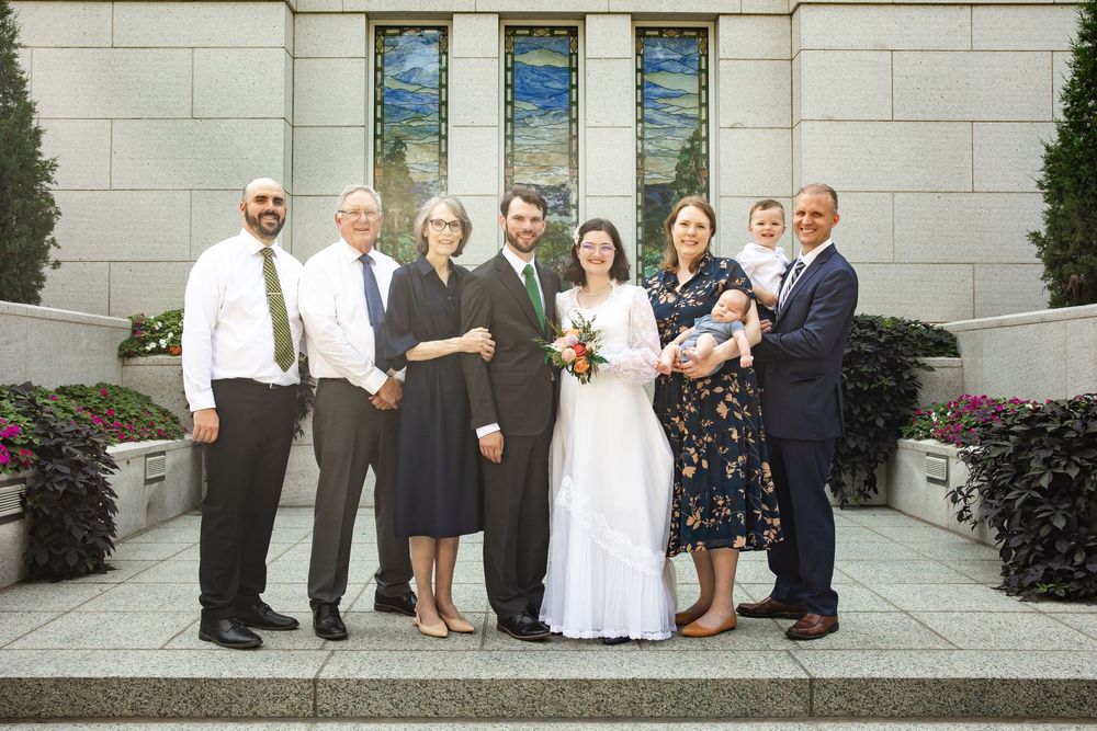 Wedding party of eight people posing outside a church, featuring bride in white gown and groom in suit.