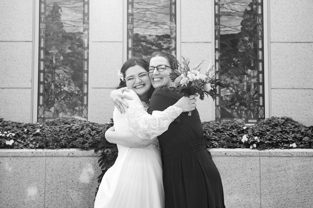 Black and white photo of a bride in white dress hugging a woman in black outside a church, both smiling joyfully.