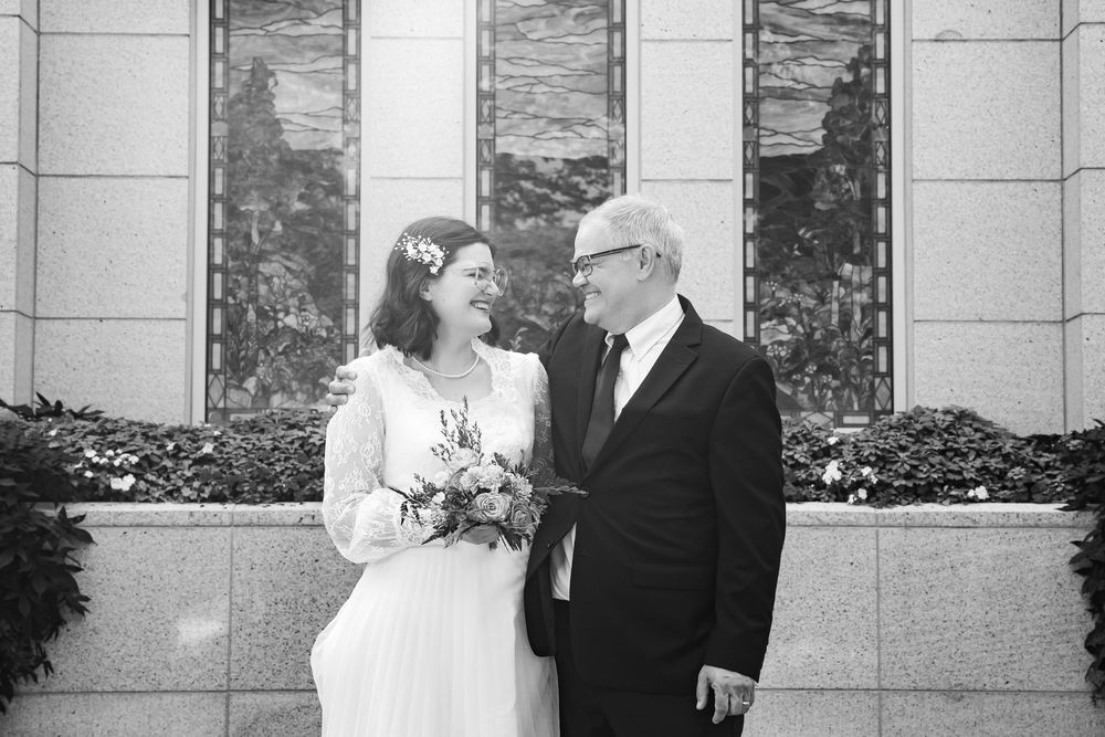 Black and white wedding photo of a smiling bride holding bouquet and groom in suit standing before ornate building.