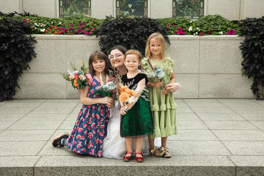 Bride posing with three young flower girls holding bouquets in front of a floral wall backdrop.