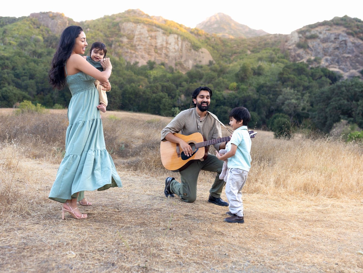 Family enjoying outdoor music session in a sunny field, father playing guitar for children while mother holds baby near scenic hills.