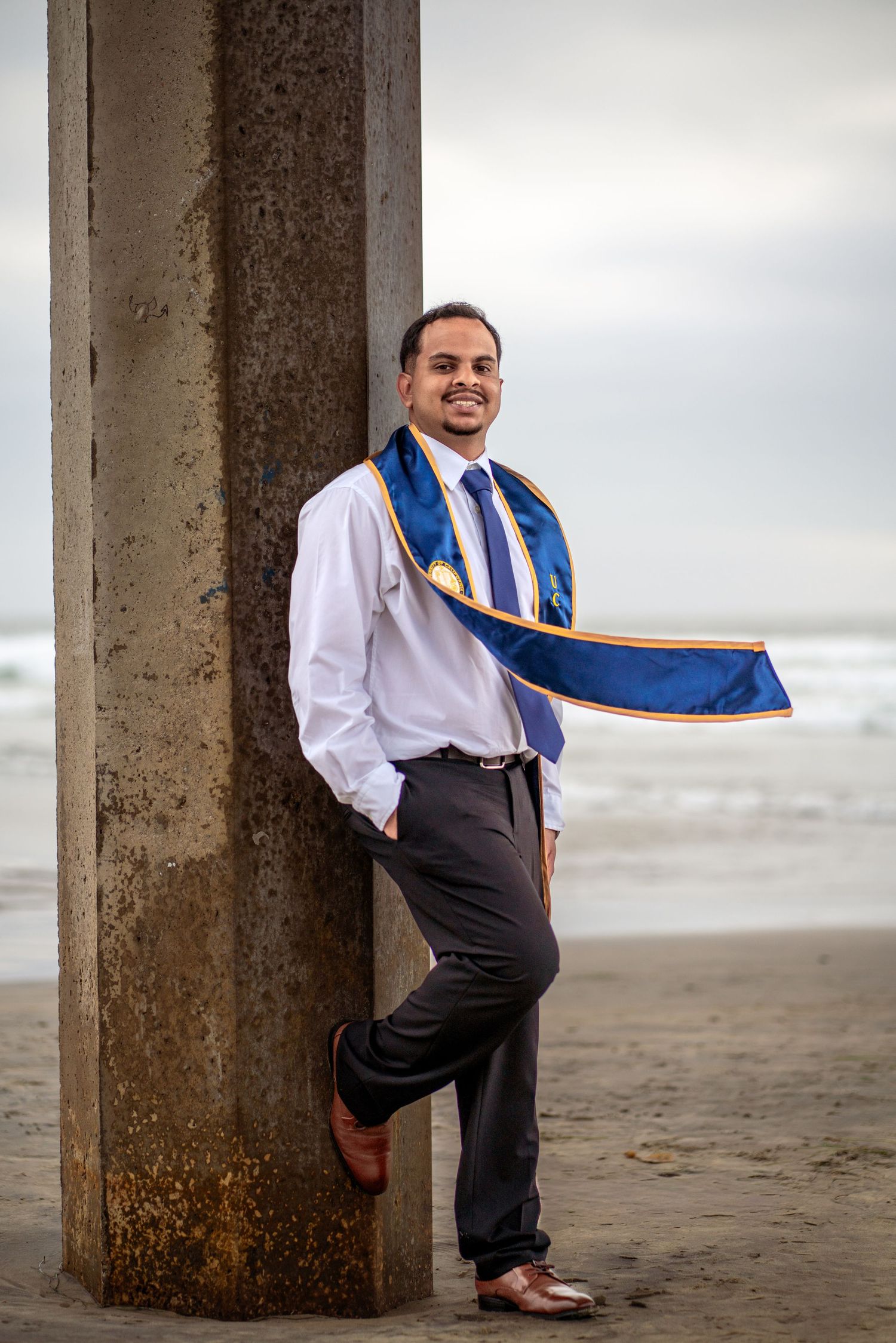 Young man in graduation sash leans against beach pier column, smiling in dress shirt and tie. La Jolla, CA