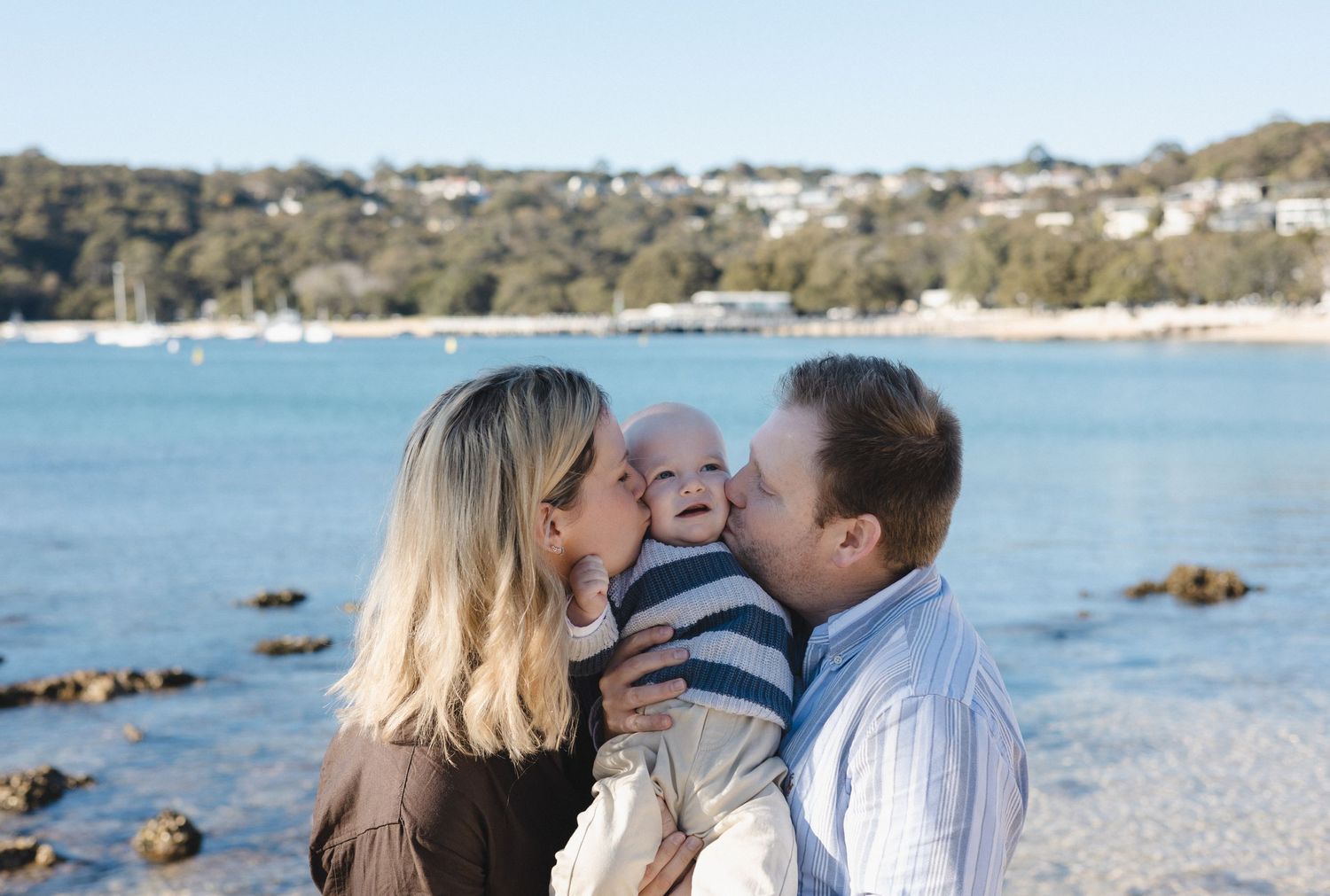Happy family with parents kissing smiling baby by Sydney harbour beach on a sunny day.