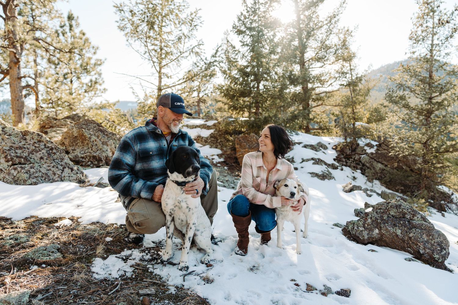Couple crouching with two dogs in snowy mountain forest with pine trees and bright sunlight.