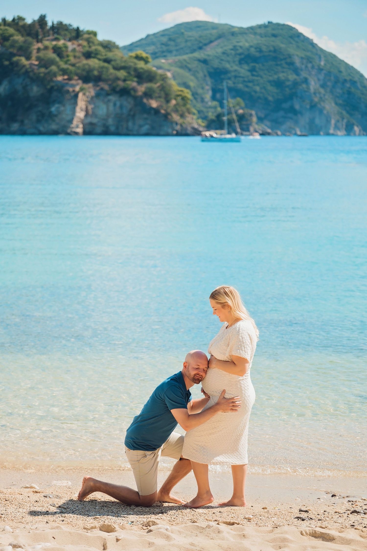 Man kneeling on beach kissing pregnant woman's belly with turquoise sea and green mountain backdrop.