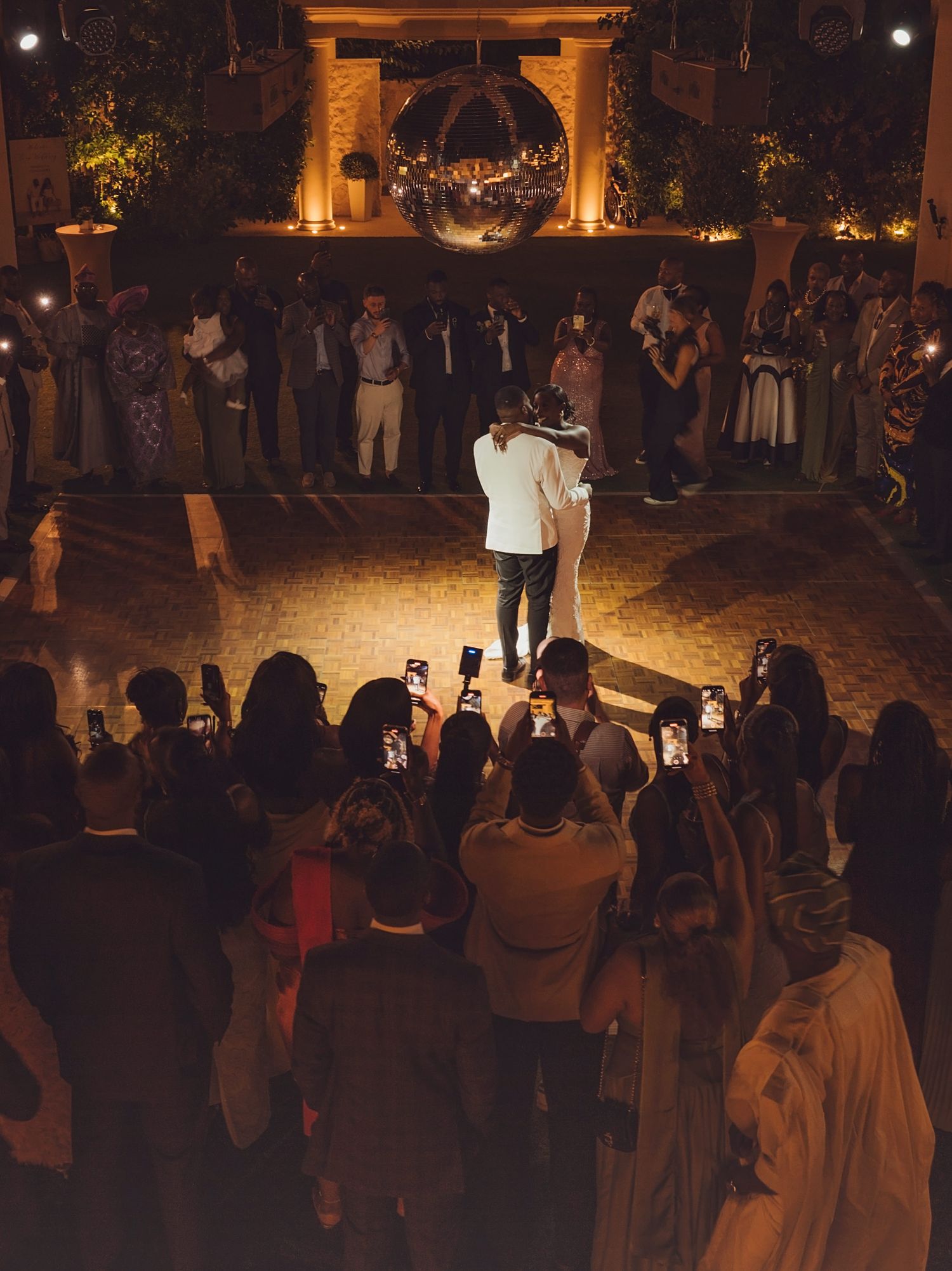 Couple shares first dance at elegant wedding reception with guests filming on phones and warm candlelit decor.