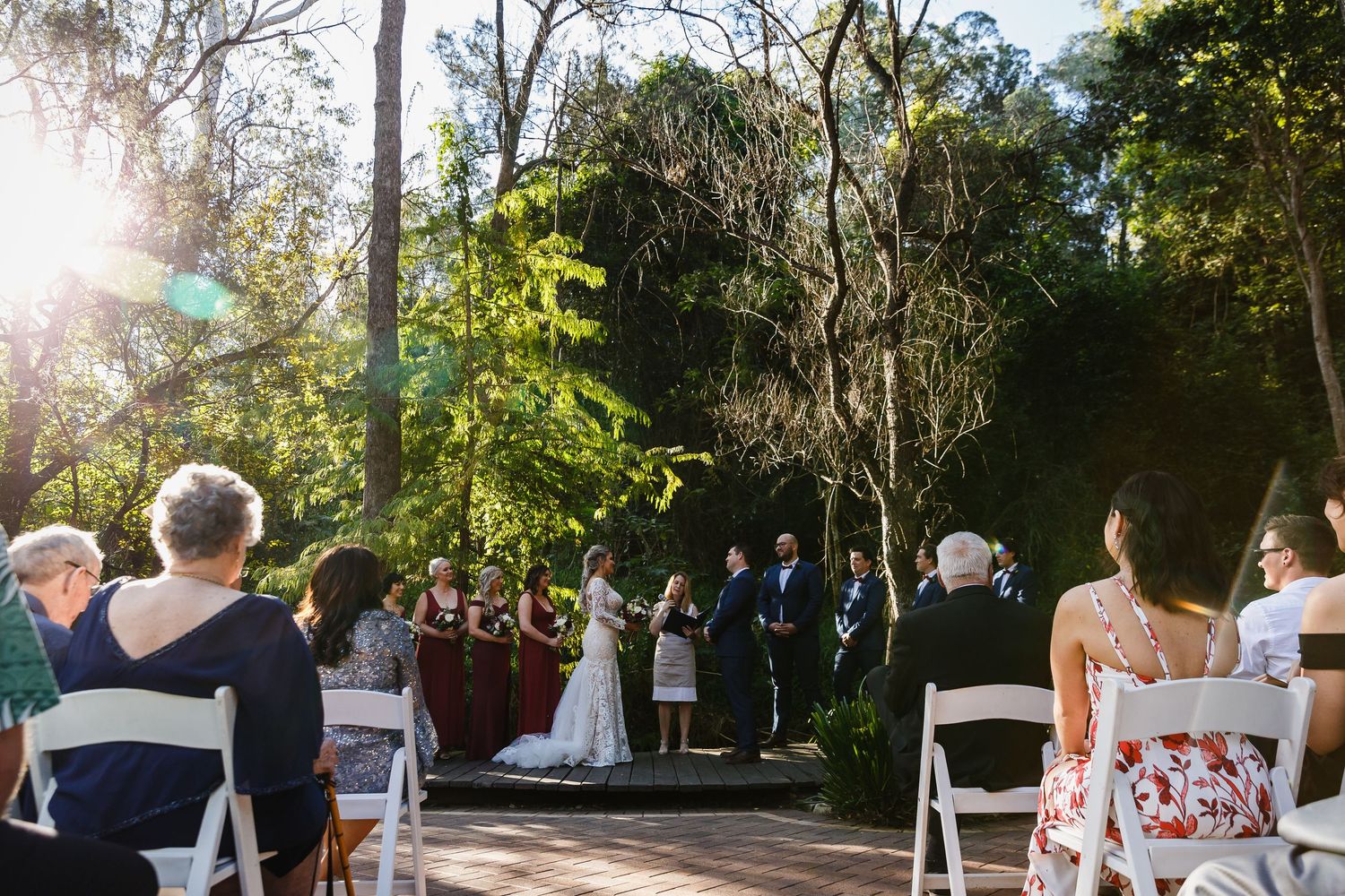 Outdoor wedding ceremony on a wooden deck surrounded by lush green trees with guests seated in white chairs at Bundaleer Rainforest Gardens