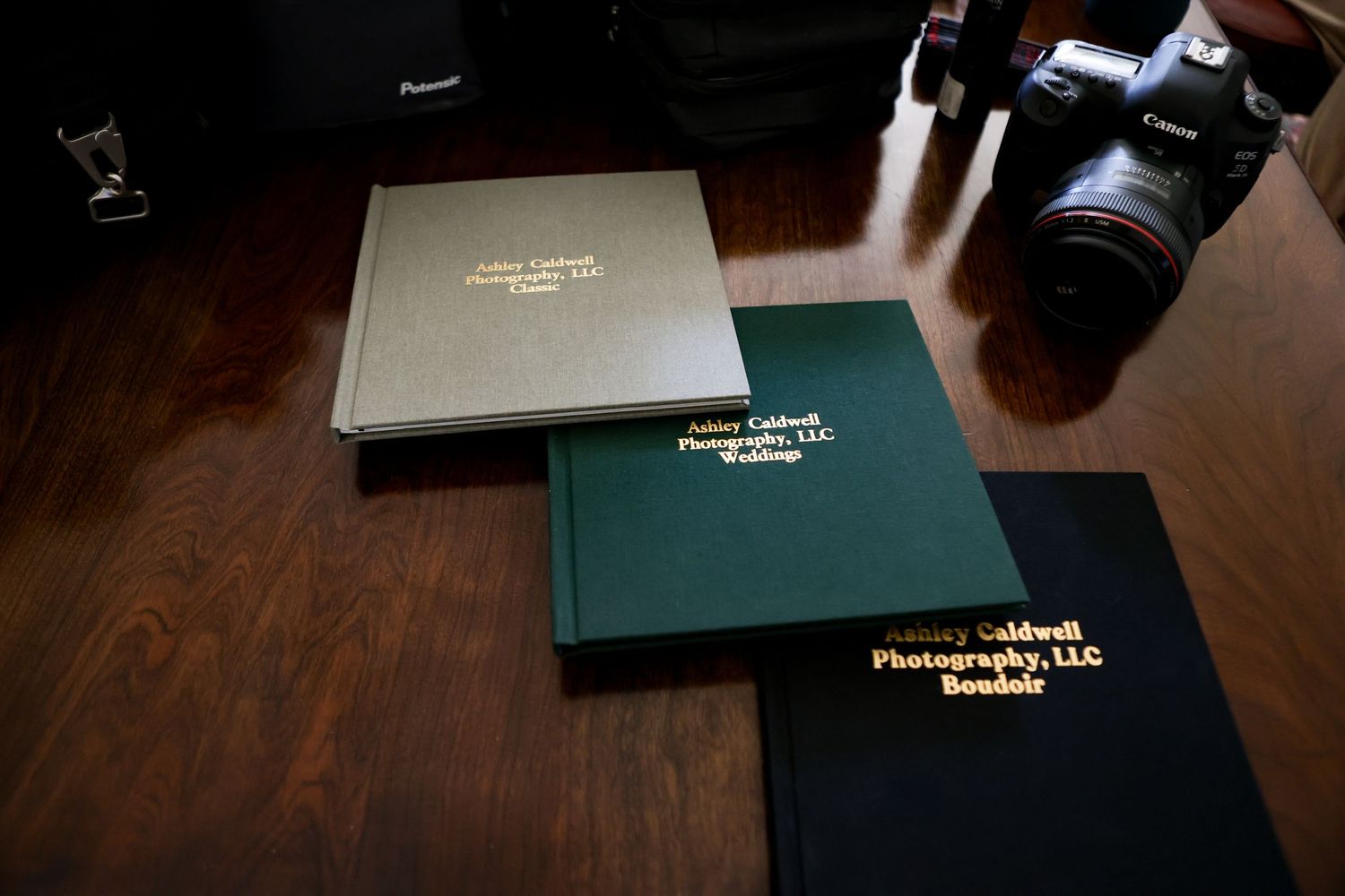 Three Ashley Caldwell Photography LLC hardcover photo books stacked on a wooden table beside a DSLR camera.
