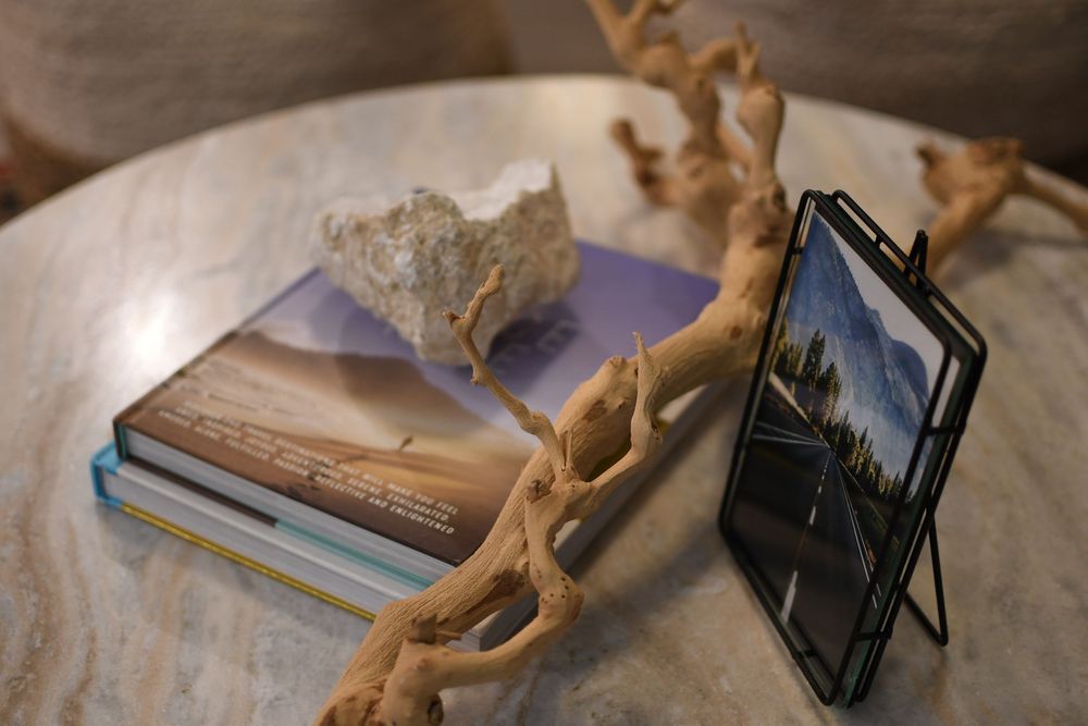Marble table with stacked books, driftwood, rock, and a tablet in a stand displaying a scenic photo.
