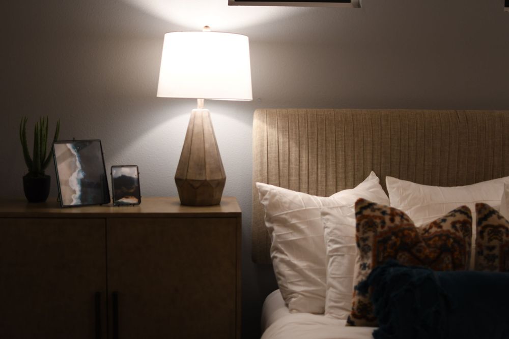 Cozy bedroom nightstand with a geometric wood lamp, framed art, and decorative plant beside a bed with white and leopard pillows.