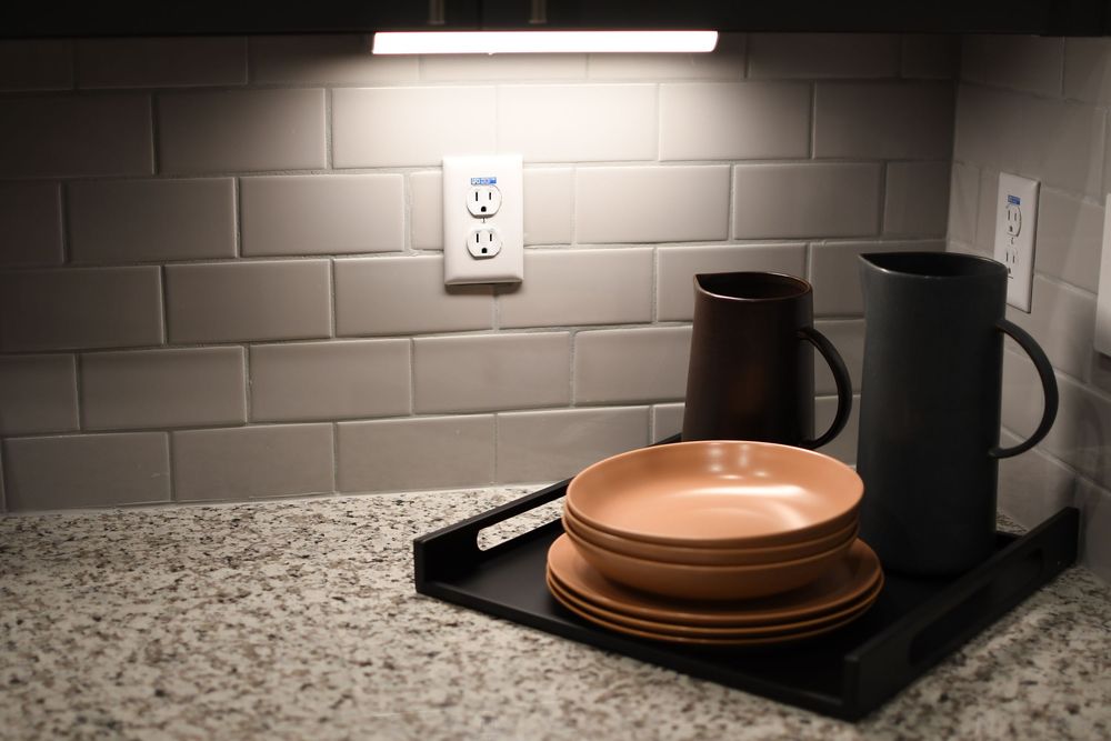 Kitchen counter with stacked terracotta dishes, two mugs on a tray, subway tile backsplash, and under-cabinet lighting.