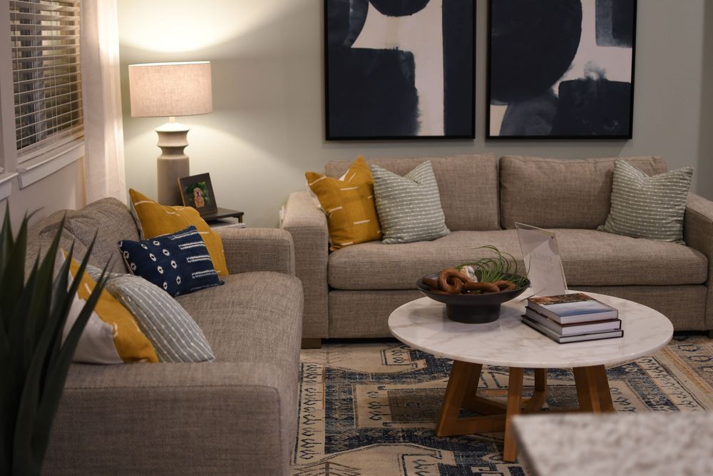 Modern living room with beige sectional sofa, yellow and blue accent pillows, marble coffee table, and abstract wall art.