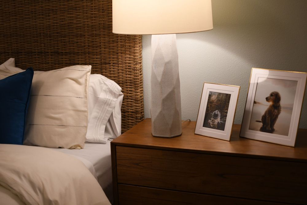 Cozy bedroom nightstand with geometric white lamp, framed dog photos, and wicker headboard with blue and white pillows.