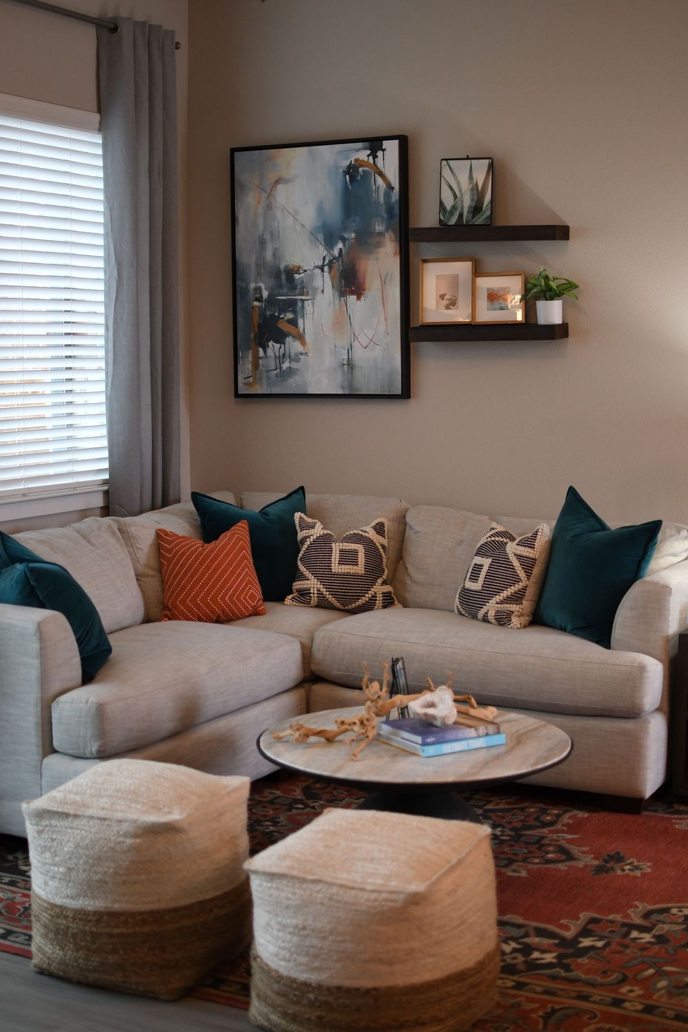 Cozy living room with beige sectional, teal and orange pillows, round coffee table, woven ottomans, and abstract wall art.