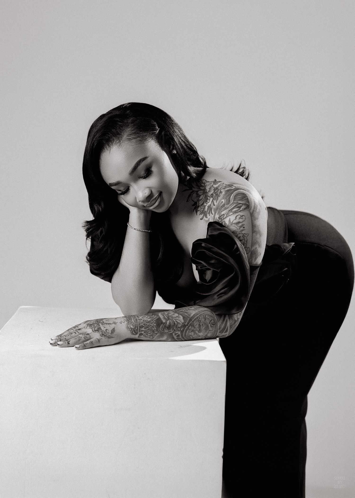 Black and white photo of a tattooed woman in elegant attire leaning gracefully on a white surface, smiling softly.