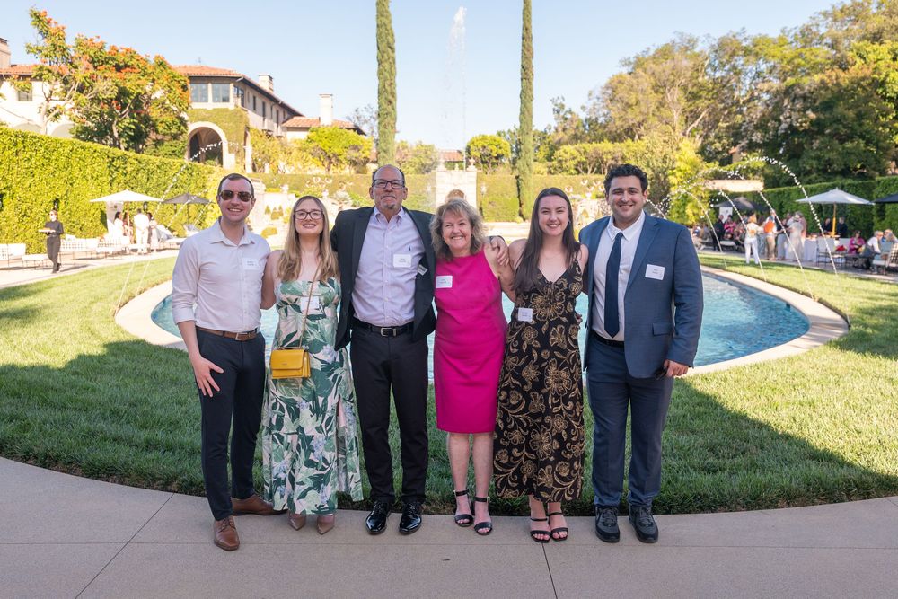 Six people pose together outdoors at an elegant garden event with a fountain and manicured lawn in the background.