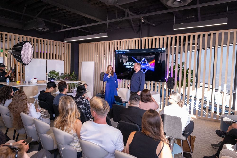 Two presenters stand before a TV screen during a corporate event presentation in a modern office space.