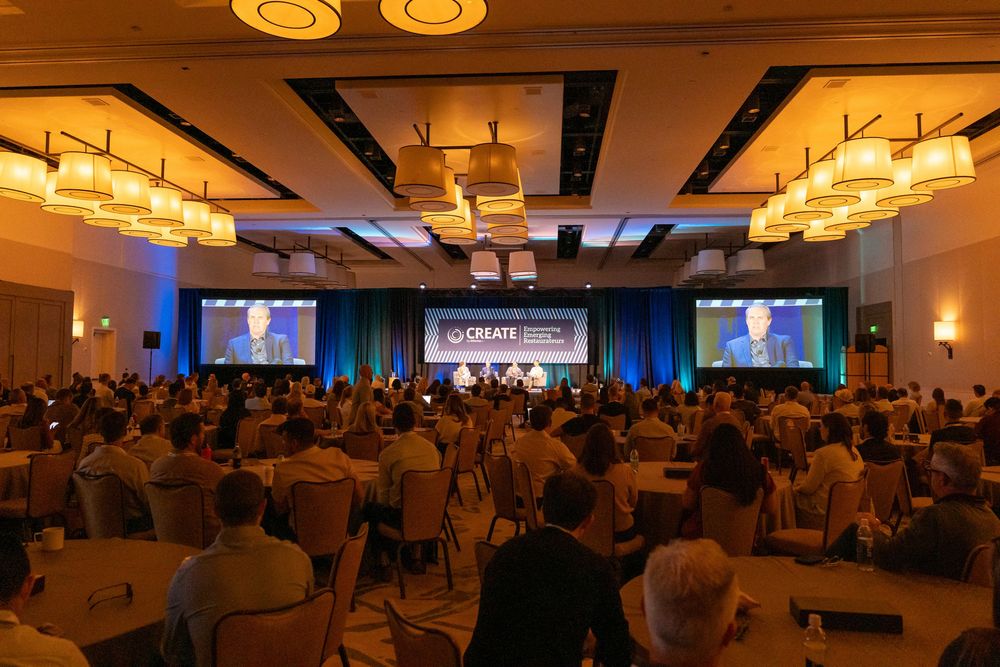 Large conference ballroom with attendees seated at tables facing a stage with two large screens displaying a speaker.