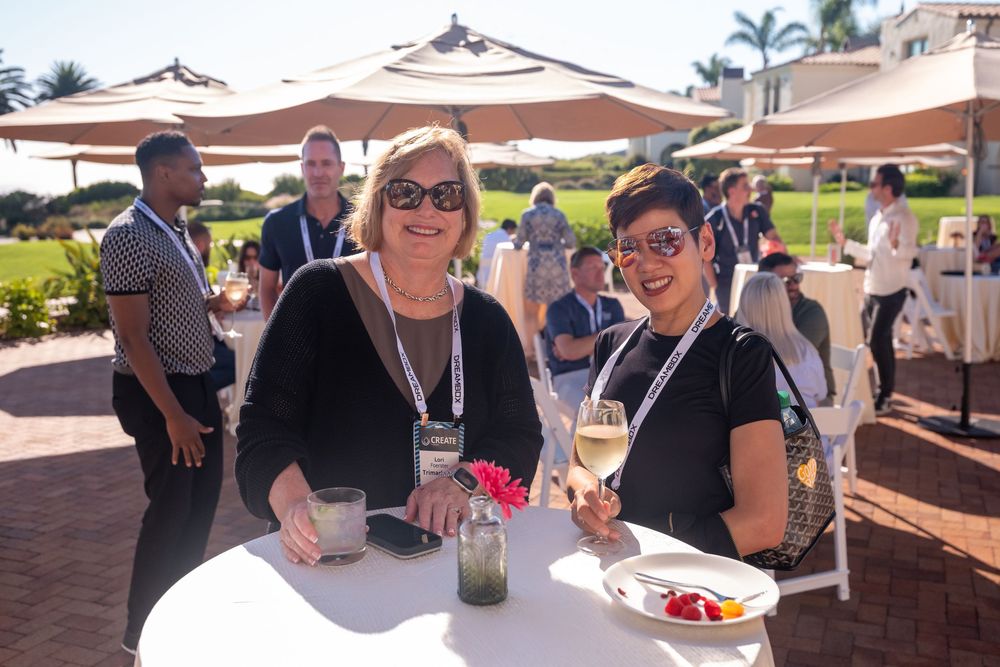Two women smile and hold drinks at an outdoor conference networking event with white umbrellas and green lawn.