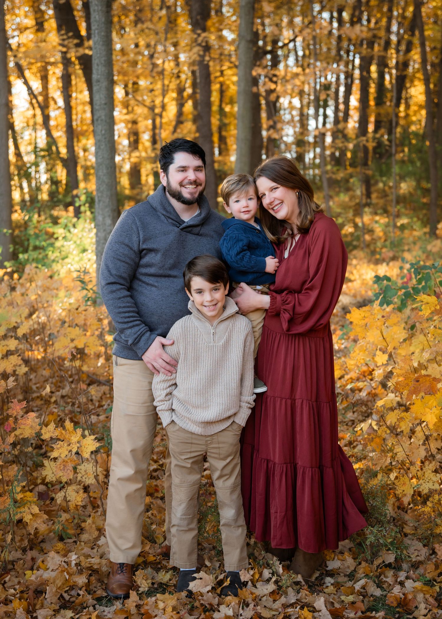Family of four smiling together in autumn forest with golden leaves, wearing casual fall outfits.