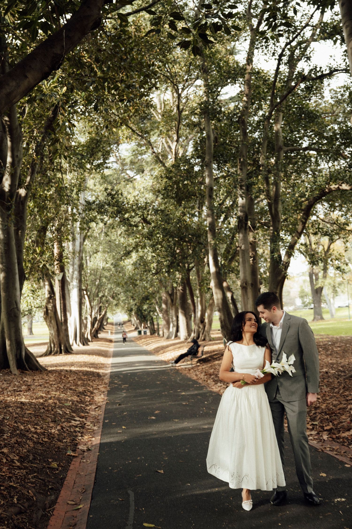 A couple in wedding attire share a romantic moment on a tree-lined path in a lush green park.