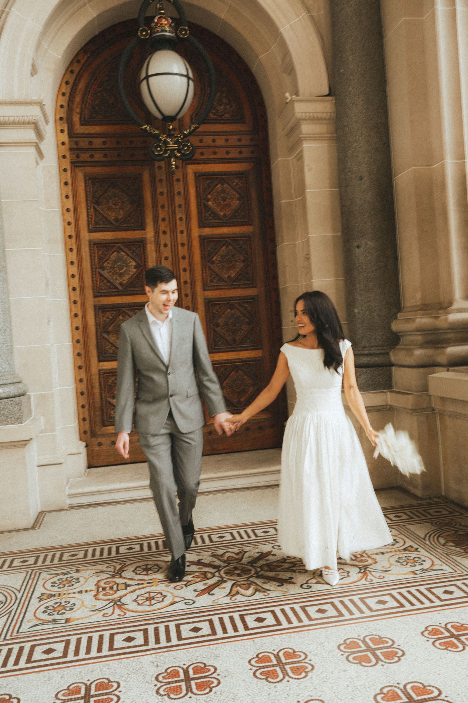 Bride and groom holding hands walking past ornate wooden doors on decorative tile floor.