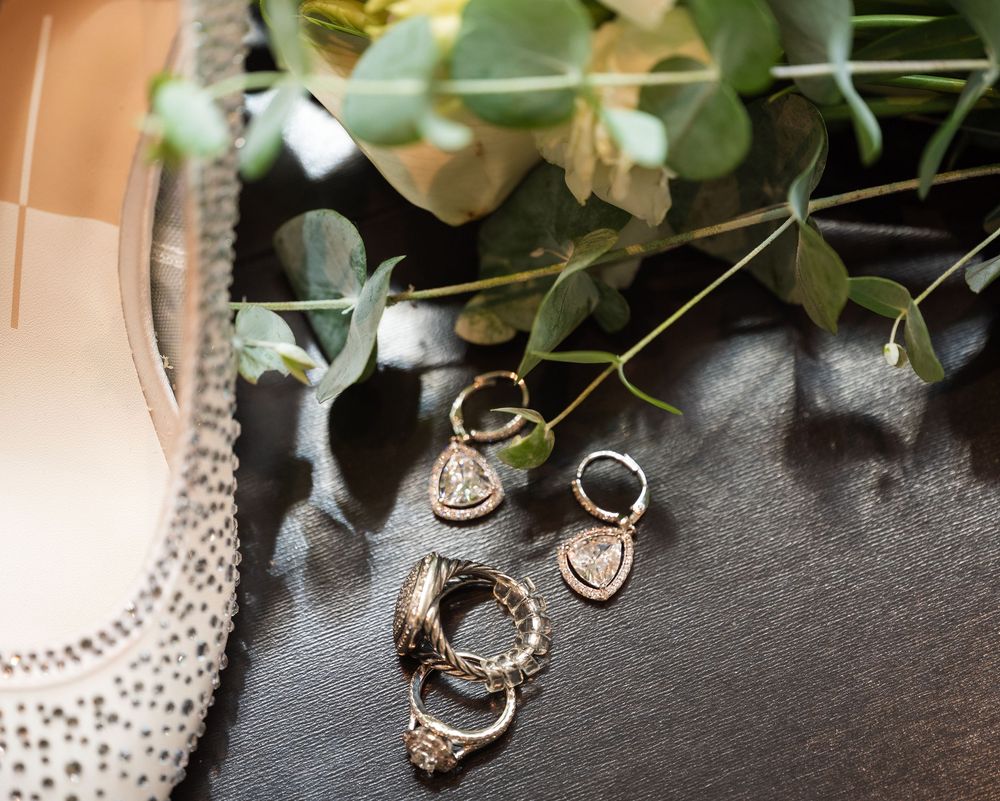 Close-up of elegant wedding jewelry including rings, earrings, and a pendant necklace arranged with greenery and white flowers.