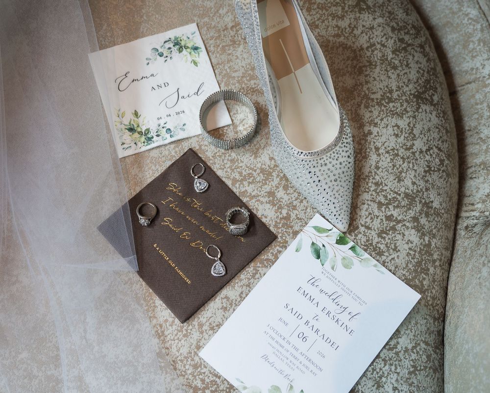 Wedding flat lay with invitation, rings, earrings, heels, and greenery stationery on elegant neutral background.