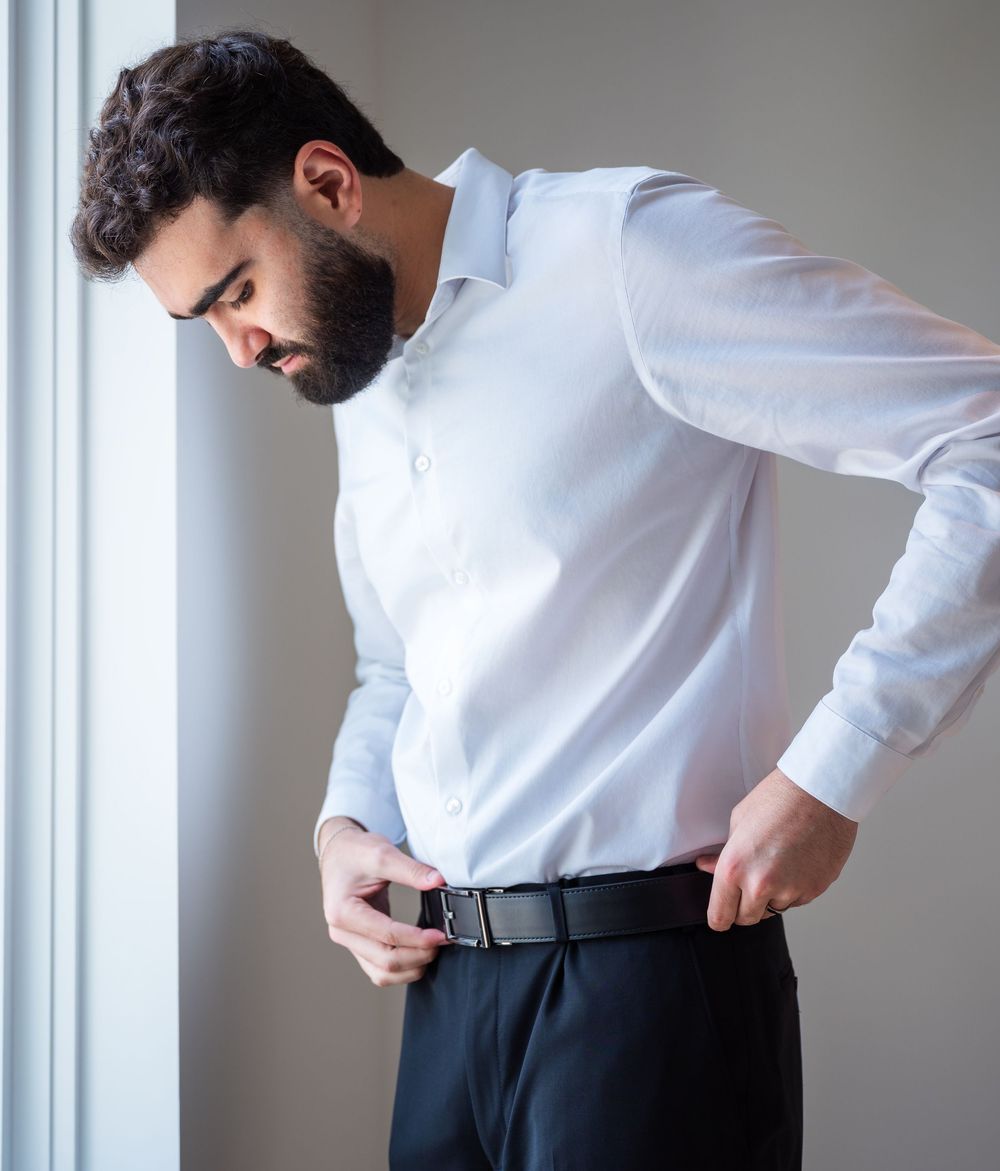 A bearded man in a white dress shirt fastening a black belt on dark trousers near a window.