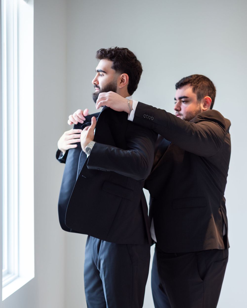 Two men in black suits, one helping the other adjust his jacket near a bright window.