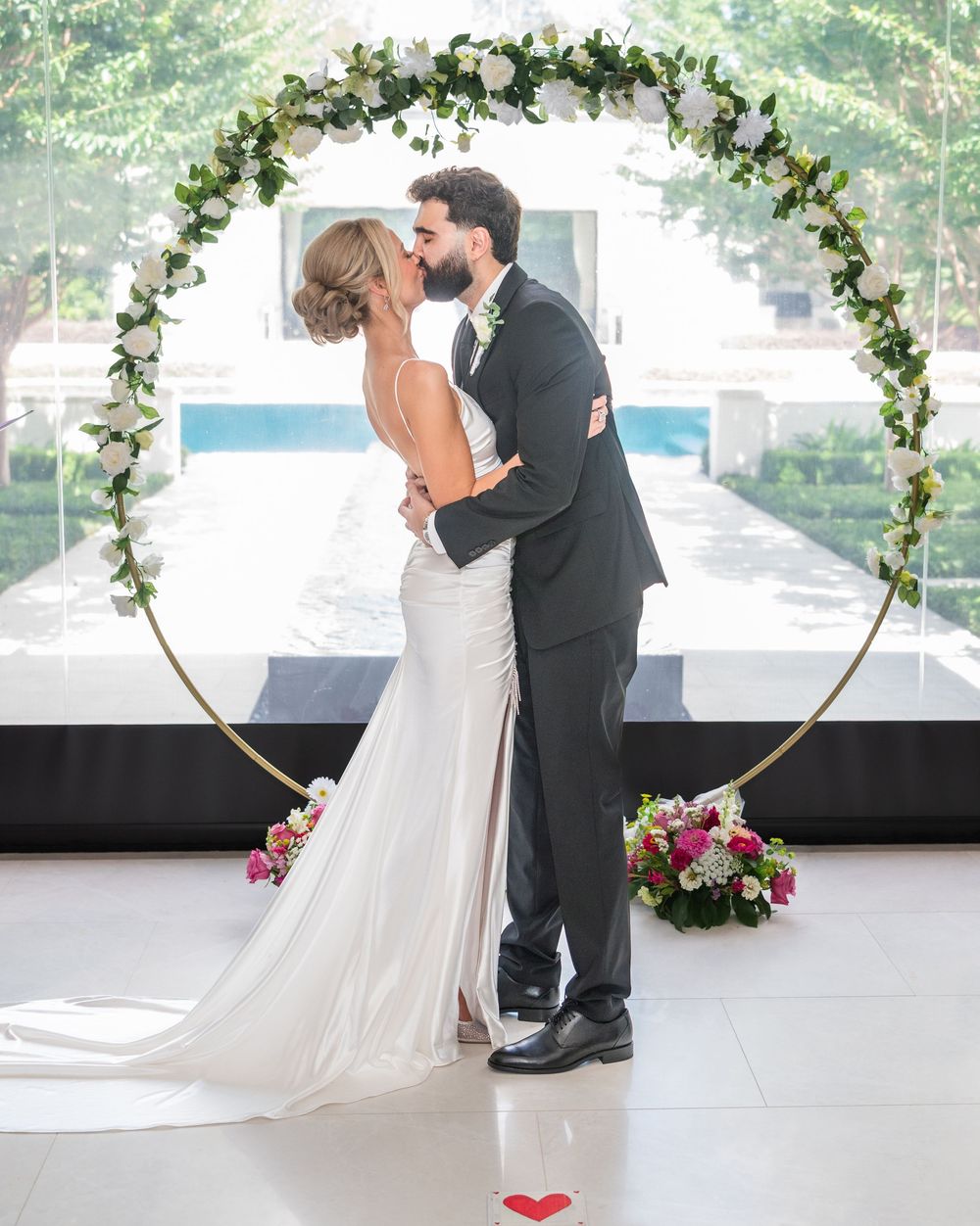 Bride and groom share first kiss under floral circle arch at elegant indoor wedding ceremony.