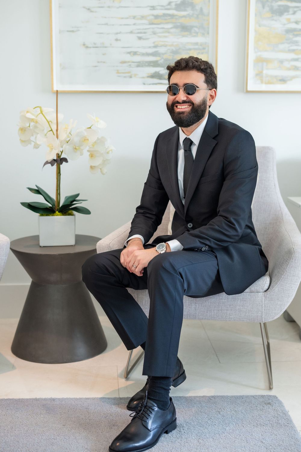 Stylish bearded man in black suit and sunglasses sitting confidently in a modern luxury interior.