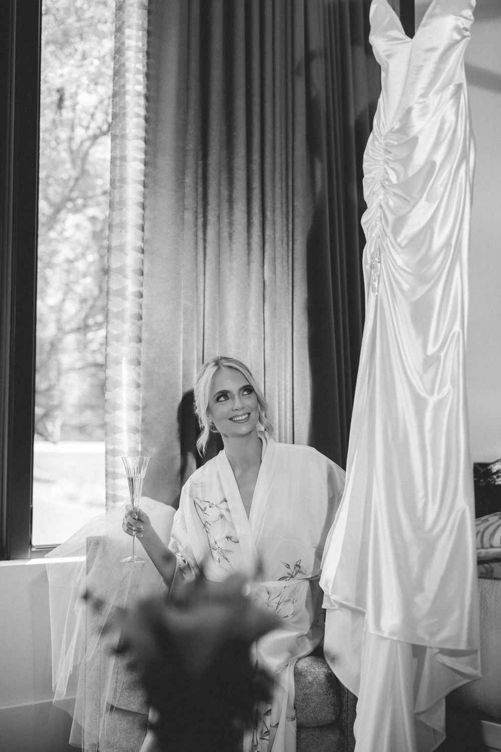 Black and white photo of a smiling bride in a floral robe holding champagne, gazing at her wedding dress hanging nearby.