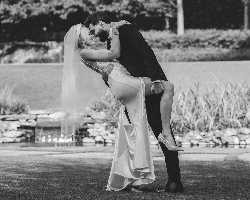 Black and white photo of a bride and groom sharing a romantic dip kiss outdoors near a garden pond.