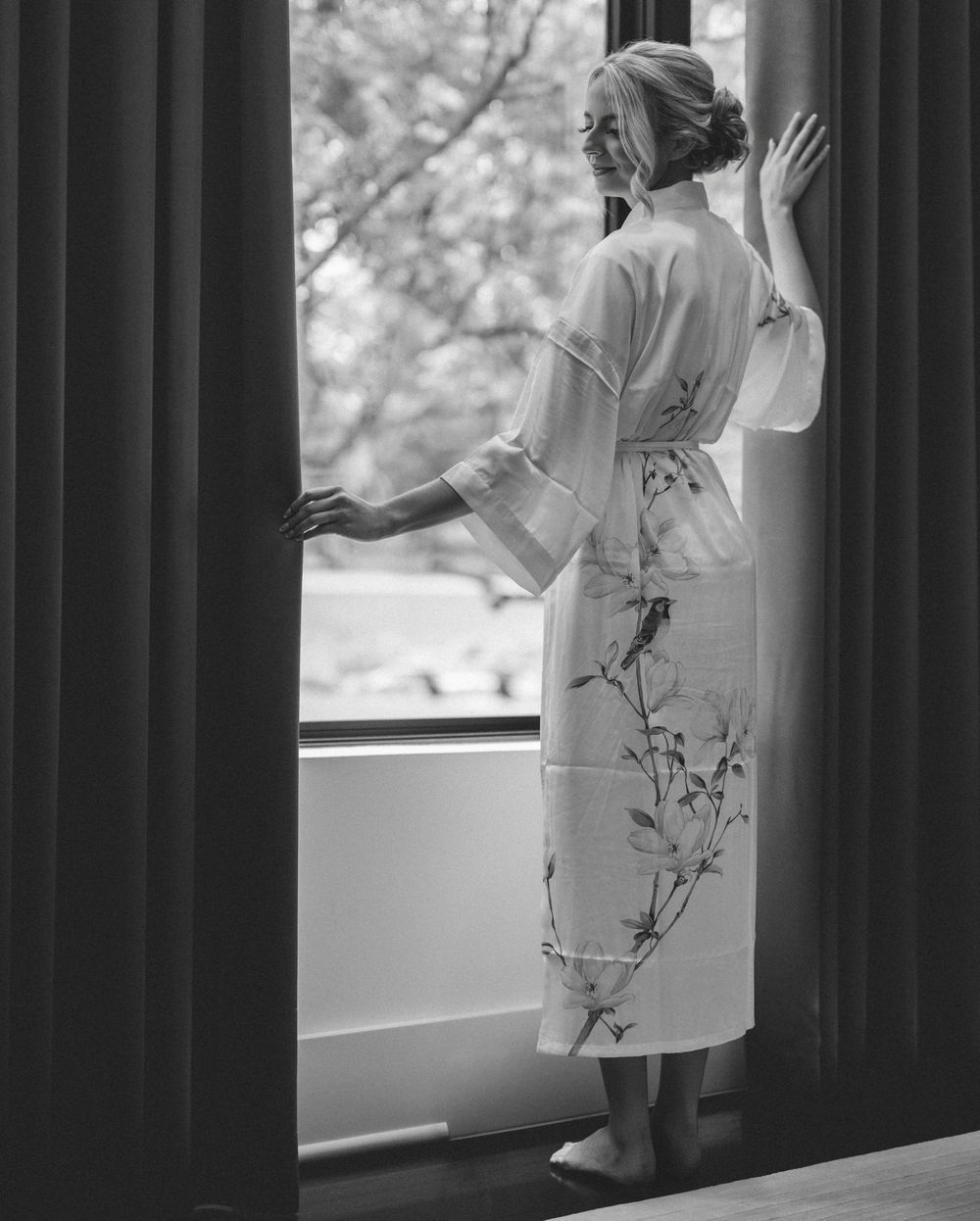 Black and white photo of a bride in a floral robe standing by a window on her wedding morning.