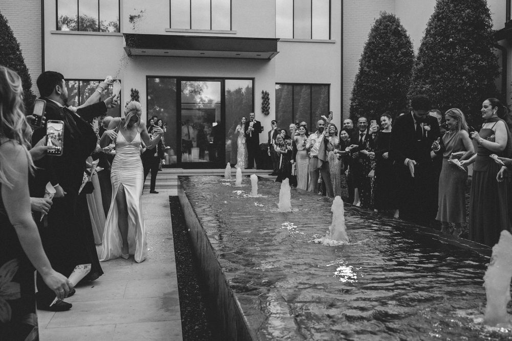 Black and white wedding photo of bride and groom walking between guests by a fountain at an elegant outdoor venue.