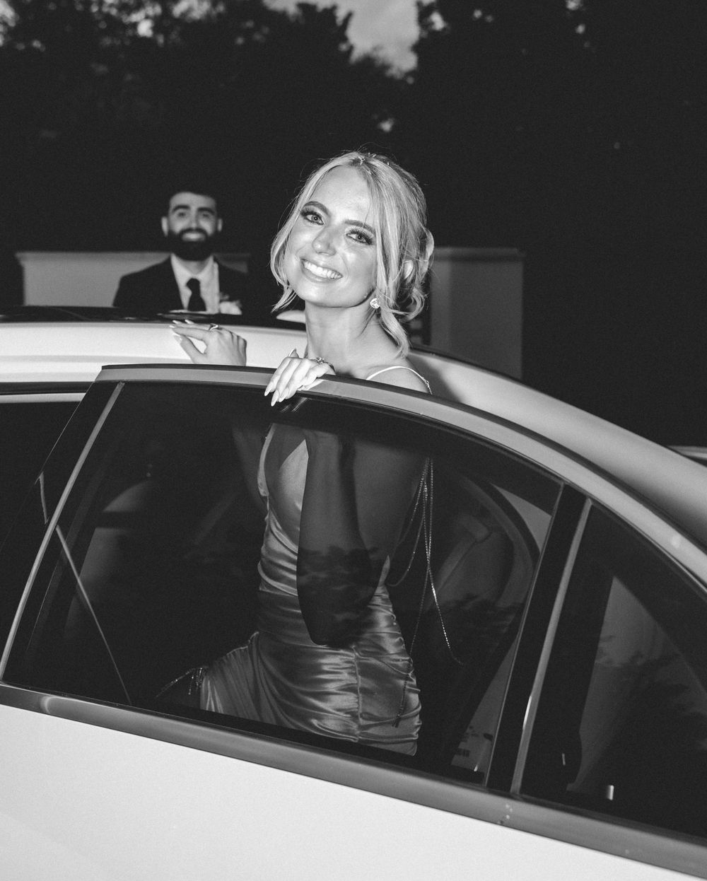 Black and white photo of a smiling blonde woman in elegant dress stepping out of a white car at a formal event.