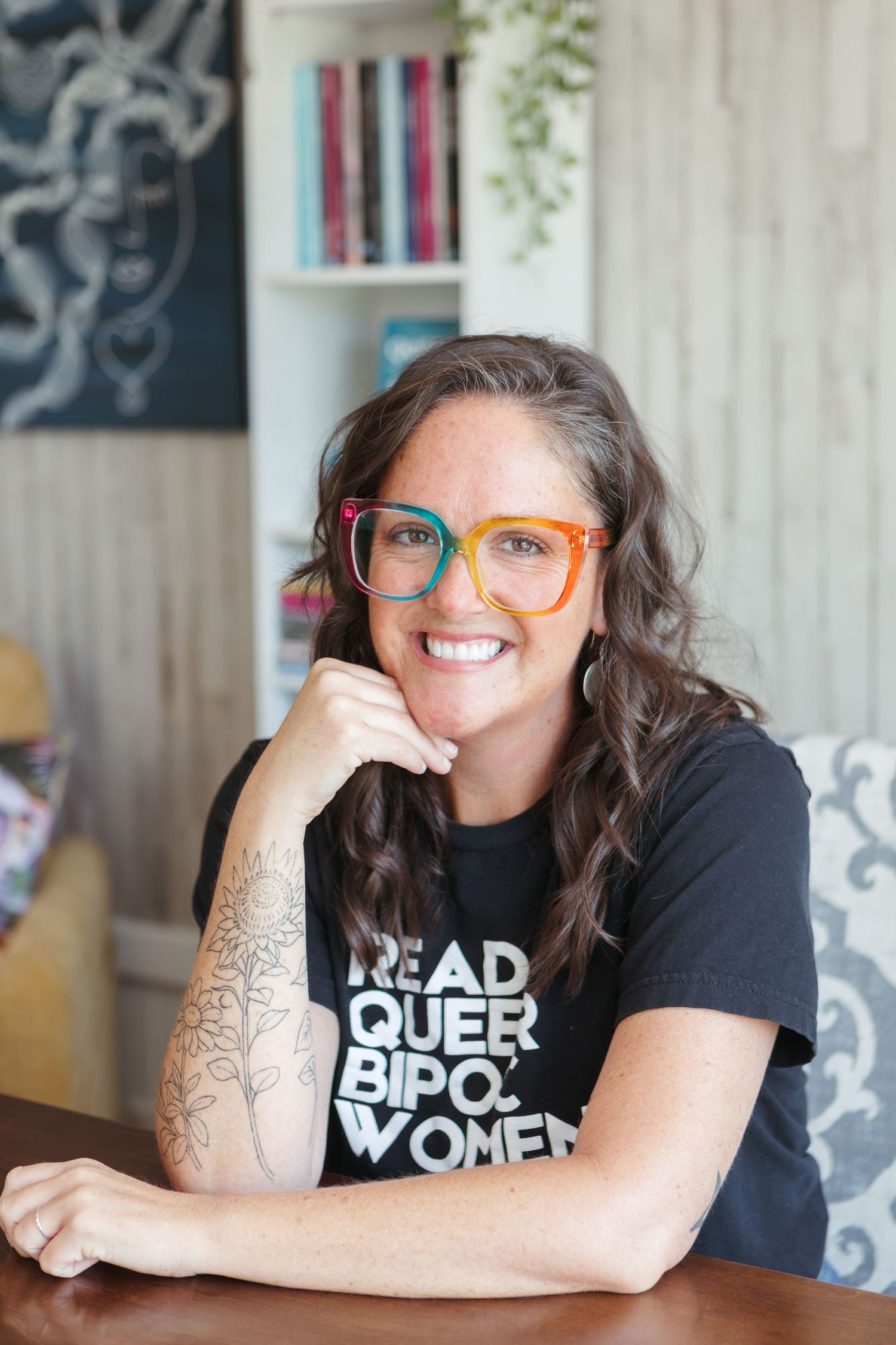 Smiling woman with colorful glasses and floral tattoo wearing a black t-shirt, seated at a wooden table indoors.