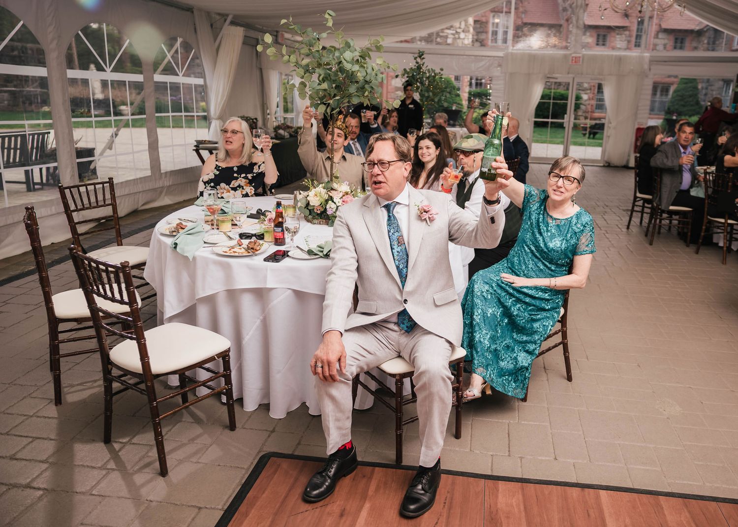 Groom's father raising a beer bottle in a toast, groom's mother raising her glass beside him.