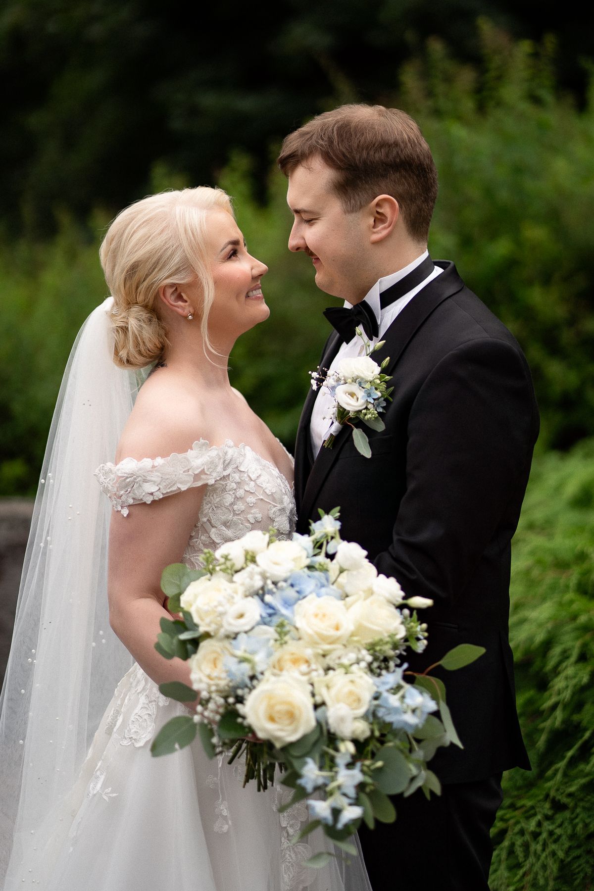 Bride in off-shoulder lace gown and groom in black tuxedo share a romantic gaze, holding white and blue bouquet outdoors.