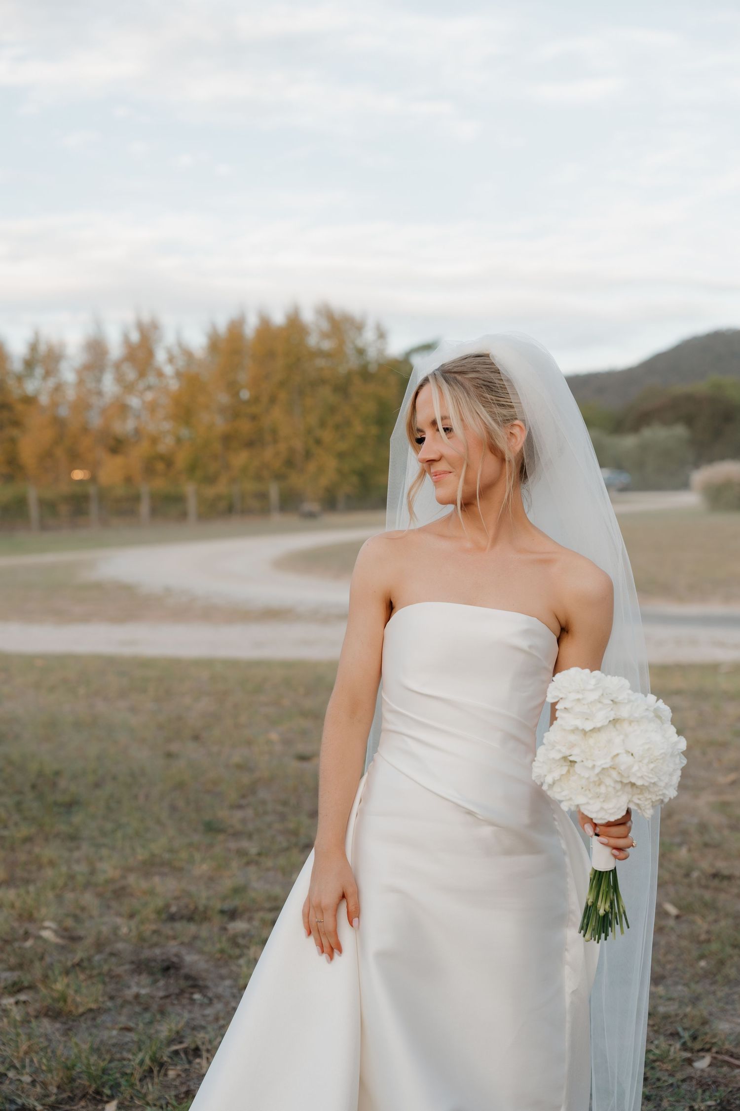 Bride in strapless white gown and veil holds white bouquet outdoors with autumn trees and hills in background.