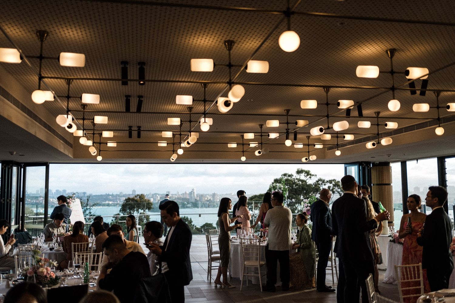 Elegant wedding reception venue with panoramic water views, guests mingling under stylish pendant lights at dusk.