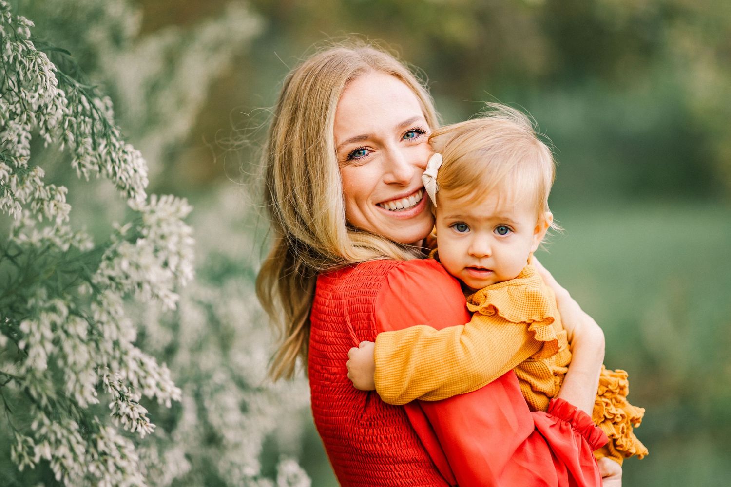 A smiling young mother in a red sweater holds her baby girl in an orange outfit outdoors among white flowers.
