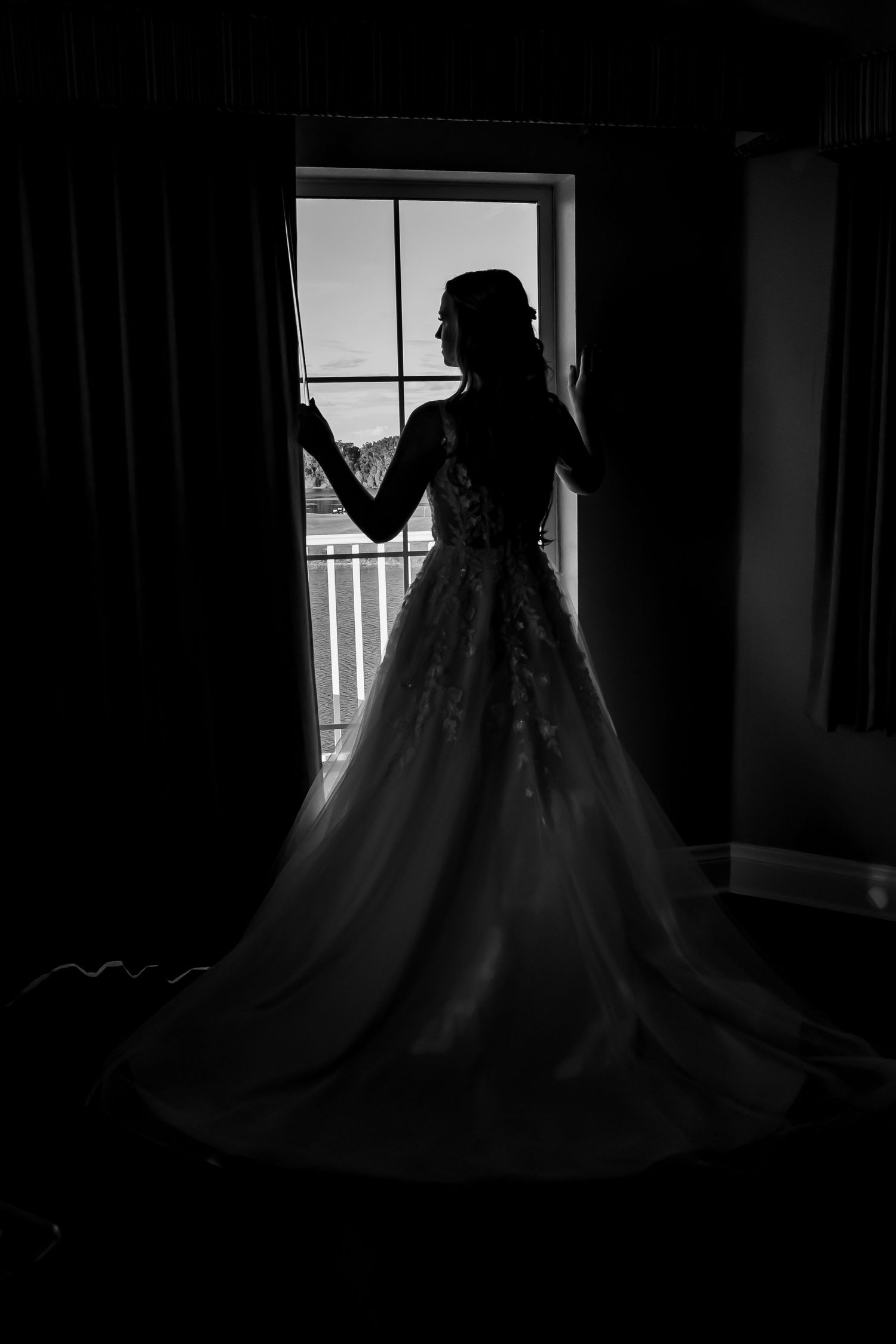 Silhouette of a bride in a flowing lace ball gown standing by a window in black and white photography.