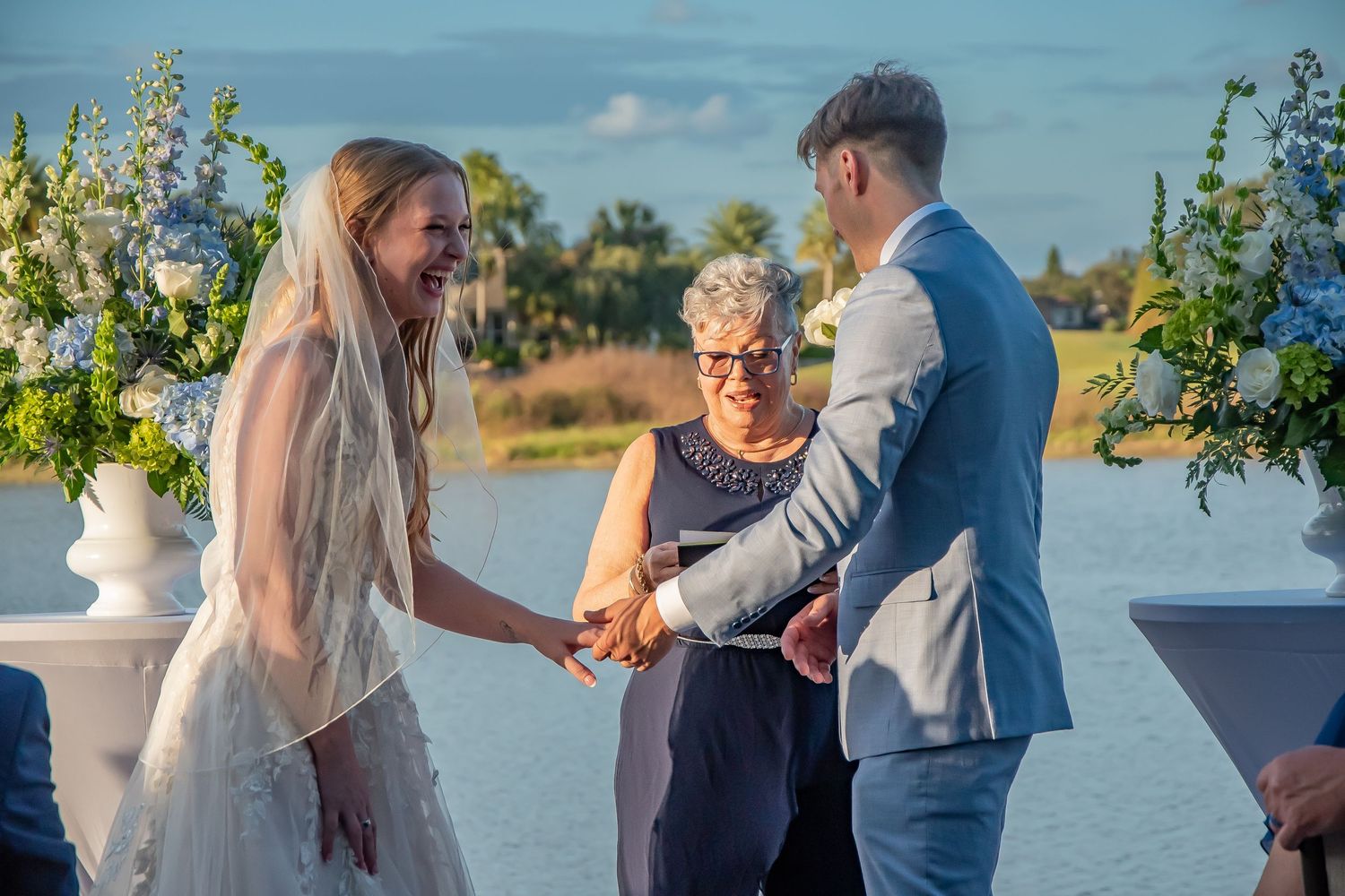 Bride and groom laughing during outdoor waterfront wedding ceremony with blue floral arrangements and officiant.