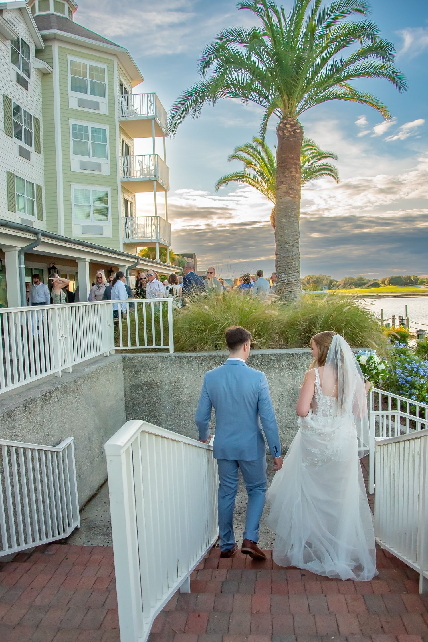Bride and groom walk hand-in-hand toward waterfront wedding reception at sunset, with palm tree and coastal hotel backdrop.