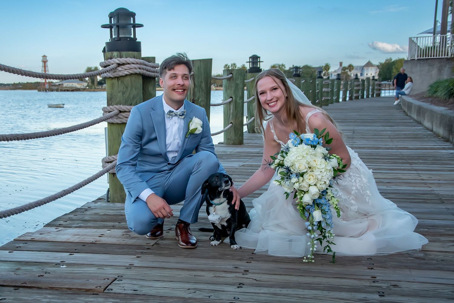 Bride and groom posing with black dog on waterfront dock, bride holding white bouquet, groom in gray suit.