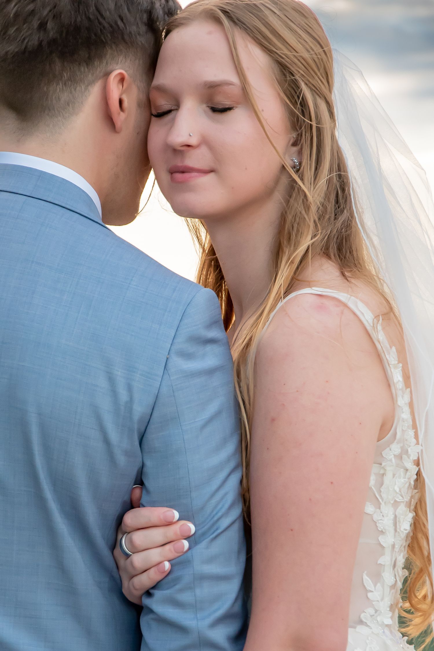 Bride in lace gown and veil embraces groom in blue suit, sharing an intimate moment with eyes closed.